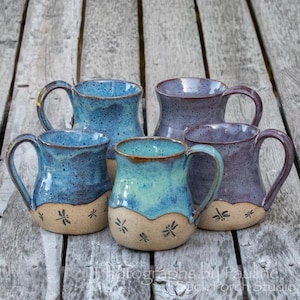 May include: Four ceramic mugs with handles, featuring a two-tone design. The upper sections are glazed in shades of blue and purple with speckled textures, while the lower sections are beige with small, dark, stamped dragonfly motifs.