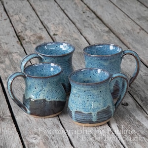 May include: Four ceramic mugs with a speckled blue glaze and dark brown base. Each mug features a rounded body, a curved handle, and a flared rim. The glaze shows variations in color intensity, with darker blue speckles throughout the surface.