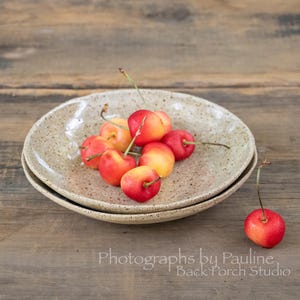 May include: A stack of two speckled ceramic plates holds a cluster of ripe cherries. The cherries are a vibrant mix of red and yellow, with green stems. One cherry rests separately on the wooden surface. The text "Photographs by Pauline Back Porch Studio" is visible.