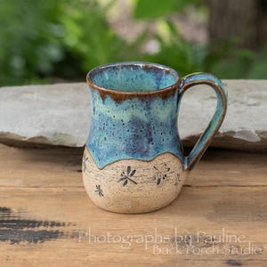 May include: A ceramic mug featuring a two-tone design with a speckled cream-colored base decorated with small black dragonfly illustrations and a glossy, dripped blue and teal glaze covering the upper portion and the handle of the vessel.