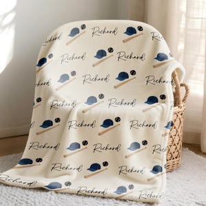 May include: A cream-coloured baseball-themed blanket featuring a repeating pattern of navy blue baseball caps, baseballs, and bats. The name "Richard" is written in a cursive font. The blanket is draped over a wicker basket.