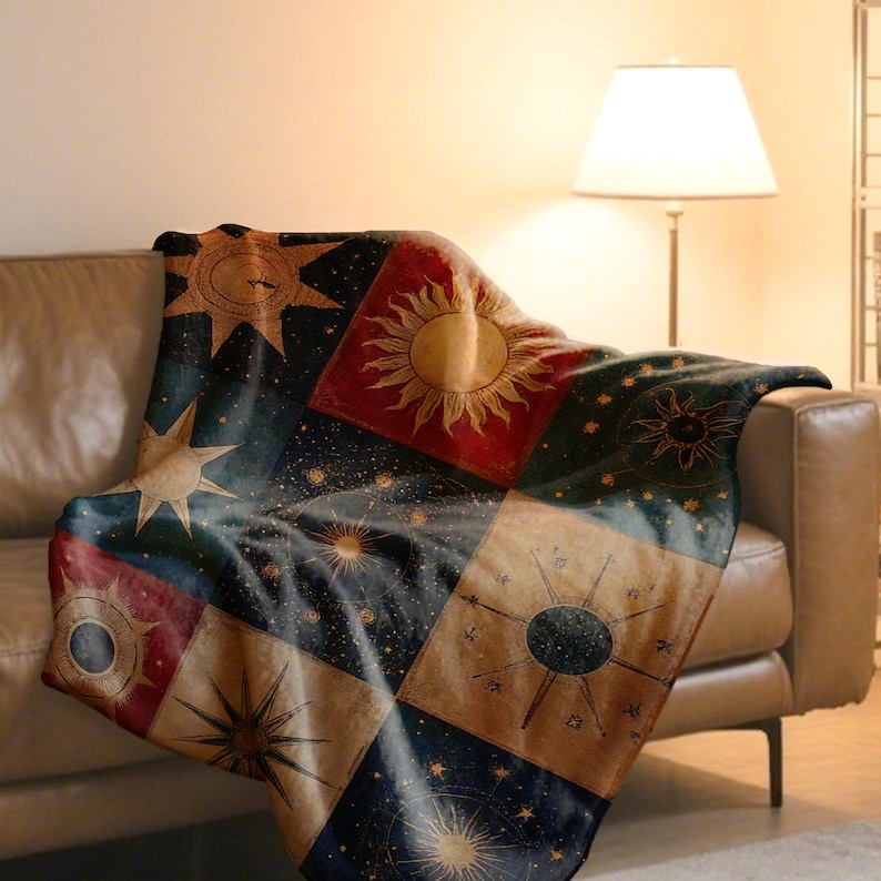 May include: A celestial-themed throw blanket with a patchwork design featuring suns, stars, and celestial patterns in shades of red, gold, and navy blue. The blanket is draped over a brown leather sofa.