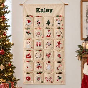Peut inclure: Un calendrier de l'Avent mural en tissu beige avec le prénom Kaley imprimé en vert foncé en haut. Il comporte vingt-quatre poches individuelles, chacune brodée d'une icône de Noël unique et numérotée de un à vingt-quatre.