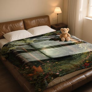 May include: A plush blanket featuring a scenic landscape design with a forest, river, and floral elements. The blanket is draped over a bed with a brown leather headboard. A teddy bear sits on the blanket.