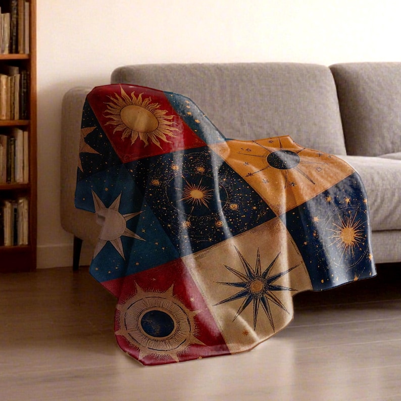 May include: A celestial-themed throw blanket draped over a gray sofa. The blanket features a patchwork design with suns, stars, and constellations in shades of red, blue, gold, and cream. The blanket is made of a soft material.