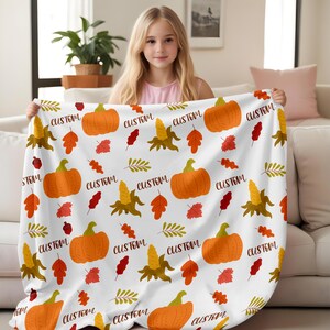 Könnte beinhalten: Weiße Fleece-Decke mit einem sich wiederholenden Muster aus orangefarbenen Kürbissen, Herbstblättern und Maisähren. Der Text "CUSTOM" wird auf der gesamten Decke wiederholt.
