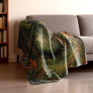 May include: A throw blanket draped over a gray sofa. The blanket features a detailed forest scene with trees, a path, and colorful flowers. The color palette includes greens, browns, and touches of red and yellow.