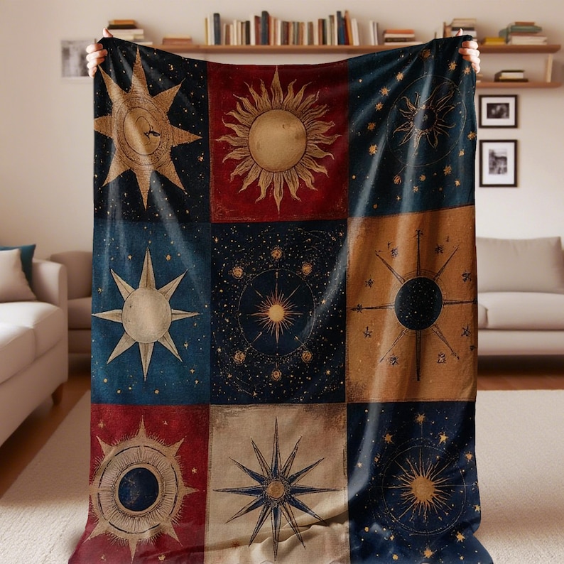 May include: A large, square blanket featuring a celestial design with suns, stars, and celestial bodies in a grid pattern. The blanket has a navy blue, red, and tan color scheme with gold and white accents. The blanket is held up by two hands.