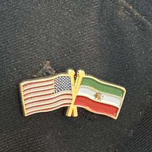 May include: A gold-coloured pin badge depicting the flags of the United States and Iran crossed. The US flag shows red and white stripes with a blue star field. The Iranian flag is green, white, and red with a central emblem.