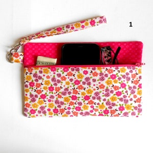 May include: A floral print wristlet with a pink zipper and wrist strap. The wristlet is filled with a black smartphone, glasses, and a small amount of cash. The floral pattern includes pink, yellow, and orange flowers on a white background.