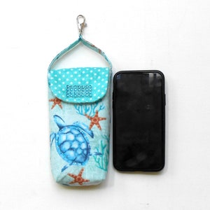 May include: A turquoise blue fabric phone case with a sea turtle design and a small pocket with rhinestones. The case has a loop for a keychain and is next to a black smartphone.