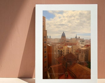 May include: A framed print of a European cityscape, featuring buildings with red tile roofs and a domed structure under a cloudy sky. The print is displayed against a peach-colored wall, with a shadow cast across the surface.