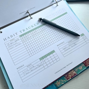 May include: A habit tracker sheet inside a teal binder with a black pen. The sheet is labeled "HABIT TRACKER" and includes sections for weekly and monthly goals. The tracker has check boxes for daily habits like walking and drinking lemon water.