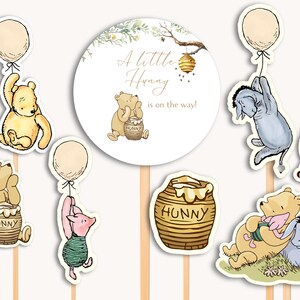 May include: Winnie the Pooh themed baby shower cake toppers. The set includes a round cake topper with the text "A little Hunny is on the way!" and several character toppers including Winnie the Pooh, Piglet, Eeyore, Tigger, and a honey pot.
