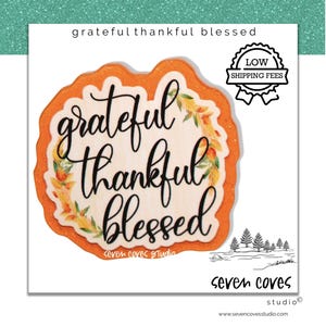 May include: A die-cut sticker featuring the text grateful thankful blessed in black cursive script. The design includes a watercolor autumn leaf border on a light wood-grain background, framed by a bright orange glittery border edge.