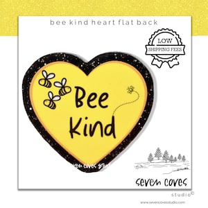 May include: A yellow heart-shaped flat back with a black glitter border, featuring the words "Bee Kind" and illustrations of bees. The text "bee kind heart flat back" is at the top. The image also includes the text "seven coves studio".