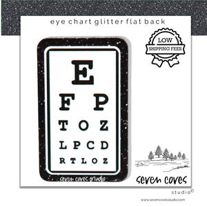 May include: A black glitter flat back with an eye chart design. The eye chart features the letters E, F, P, T, O, Z, L, C, D, and R. The text "eye chart glitter flat back" is at the top. The text "seven coves studio" is at the bottom.