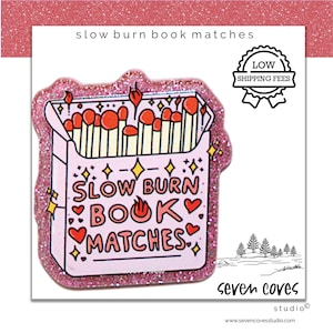 May include: A pink glittery sticker featuring a box of matches with the text "SLOW BURN BOOK MATCHES". The match heads are red, and the box has hearts and stars. The sticker also has the text "seven coves studio".