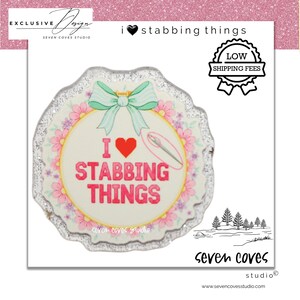 May include: A round, decorative pin with the text "I ❤️ Stabbing Things" in pink, surrounded by a floral design and a mint green bow. The pin has a silver glitter edge and the words "Seven Coves Studio" are also visible.