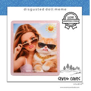 May include: A square pink glittery coaster featuring a printed image of a doll with long brown hair and sunglasses next to an orange cat wearing sunglasses and a gold chain. The text reads disgusted doll meme, seven coves studio, and low shipping fees.