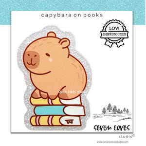 May include: A cute cartoon capybara character sitting on a stack of books. The books are yellow, teal, and pink with white pages. The image has the text "capybara on books" and the logo of "seven coves studio".