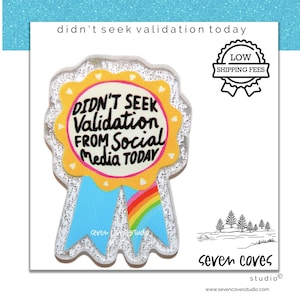 May include: A glittery award ribbon pin featuring a yellow center with the black text DIDN'T SEEK VALIDATION FROM SOCIAL MEDIA TODAY. The ribbon has two light blue tails, one of which displays a colorful rainbow graphic, all encased in a clear, sparkling acrylic border.
