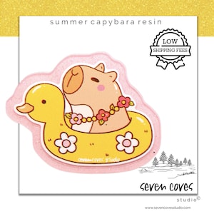 May include: A resin sticker featuring a cartoon capybara wearing a flower lei and floating in a yellow duck-shaped pool float. The sticker has the text "summer capybara resin" and the logo "seven coves studio".