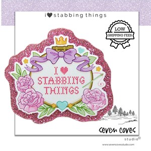 May include: A glittery pink die-cut sticker featuring an embroidery hoop illustration with the text I heart STABBING THINGS in a cross-stitch font. The design includes a purple bow, a gold crown, pink roses, green leaves, and a teal heart.