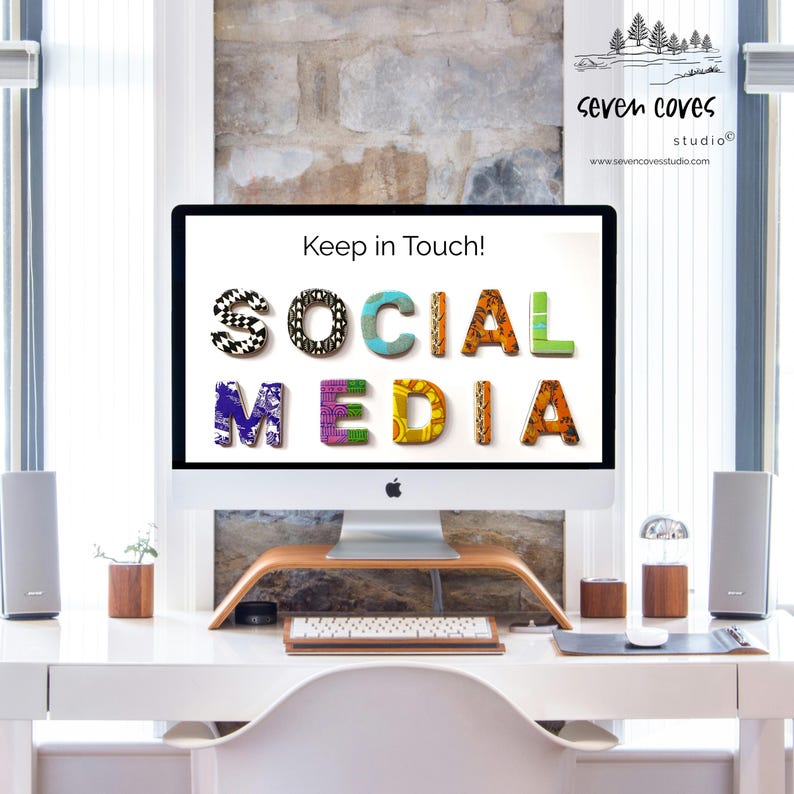 May include: A desktop computer screen shows the text "Keep in Touch!" and "SOCIAL MEDIA" in vibrant, patterned lettering. The computer is on a white desk with speakers, a keyboard, and a wooden monitor stand. A brick wall forms the backdrop.