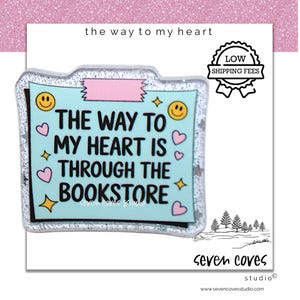 May include: A light blue sticker with the text "THE WAY TO MY HEART IS THROUGH THE BOOKSTORE". The sticker has a glittery border, pink tape, and features smiley faces, hearts, and stars. The sticker is on a white background with a pink glitter top.
