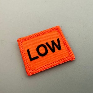 Könnte beinhalten: Orangener Stoff-Patch mit schwarzen Nähten und dem Wort "LOW" in schwarzen Buchstaben.