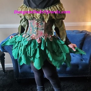 May include: A fantasy-themed costume featuring a green leaf-like skirt and bodice with floral accents. The top has a corset-style design with lace-up detailing. The sleeves are olive green, and the outfit is paired with gray leggings and socks. The website DungeonDudsJewels.com is visible.