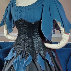 May include: A fantasy-style dress with a blue bodice and sleeves, a black corset, and a layered skirt. The skirt features blue and black petal-like accents over a white underskirt. The corset has a lace-up back. The website address "dungeondudsnjewels.com" is visible.