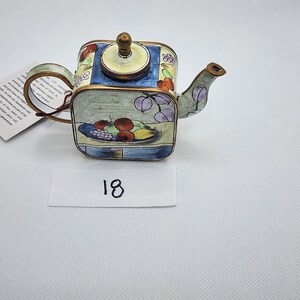 May include: A miniature decorative teapot with a square body and a curved spout. The teapot features a painted design of fruit on a plate, with a blue and yellow background. The handle and spout are a bronze color. The number "18" is written on a white label.