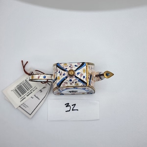 May include: A small, white teapot with blue and gold floral designs. The teapot has a spout and a handle. The teapot is sitting on a white surface.