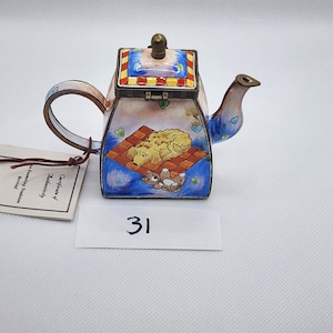 May include: A small, blue and white enamel teapot with a red and yellow checkered lid. The teapot features a painted scene of a dog and a teddy bear on a blue blanket. The teapot is labeled with the number "31".