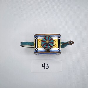 May include: A small, colorful teapot with a blue, yellow, red, and green design. The teapot has a round lid with a green and red design. The teapot is sitting on a white surface. The number 43 is written on a white label below the teapot.