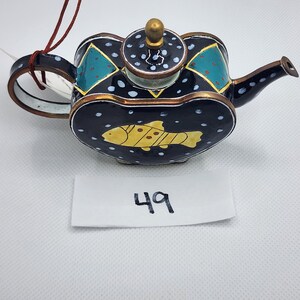 May include: A small, heart-shaped teapot with a black base and white polka dots. The teapot has a gold trim and a yellow fish painted on the front. The teapot has a green and blue design on the top and a gold lid.