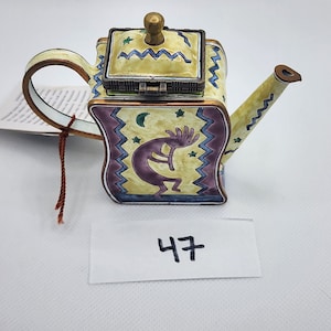 May include: Decorative miniature teapot with a hinged lid, featuring a Kokopelli figure. The teapot has a yellow background with blue and purple accents, and a gold-colored handle and spout. The number "47" is written on a white label.