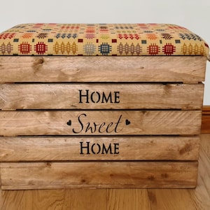 May include: A wooden storage box with a cushioned top, featuring a colourful patterned fabric. The box has rope handles and is labelled "Home Sweet Home".