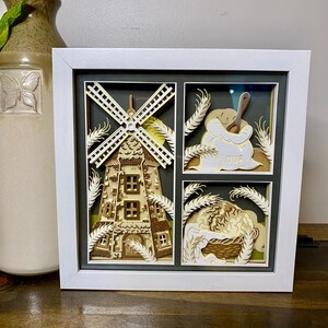 May include: A framed paper art piece featuring a windmill, a bag of flour, and a basket of bread. The artwork is multi-layered with a white frame and a dark green background. Wheat stalks are also included in the design.