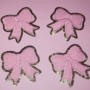 May include: Four pink felt bows with gold glitter trim. The bows are on a pink background.