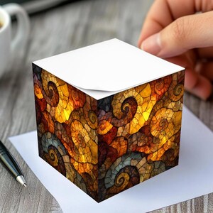 May include: A cube-shaped notepad with a colorful, abstract design of swirling patterns in shades of gold, orange, and brown. The notepad is on a white sheet of paper, with a pen and a cup of coffee in the background.