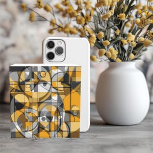 May include: A white smartphone case stands next to a decorative cube with a geometric pattern in shades of yellow, gray, and black. A white vase holds yellow flowers, adding a pop of color to the scene. The cube's design features circles and squares.