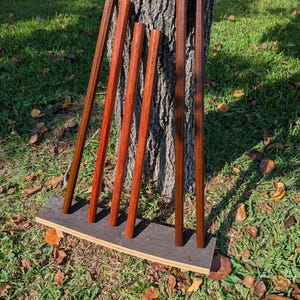 May include: Six wooden dowels of varying shades of brown and red are arranged vertically, leaning against a tree trunk. They are set in a dark gray base, resting on a bed of grass and fallen leaves. The dowels appear to be of different wood types.