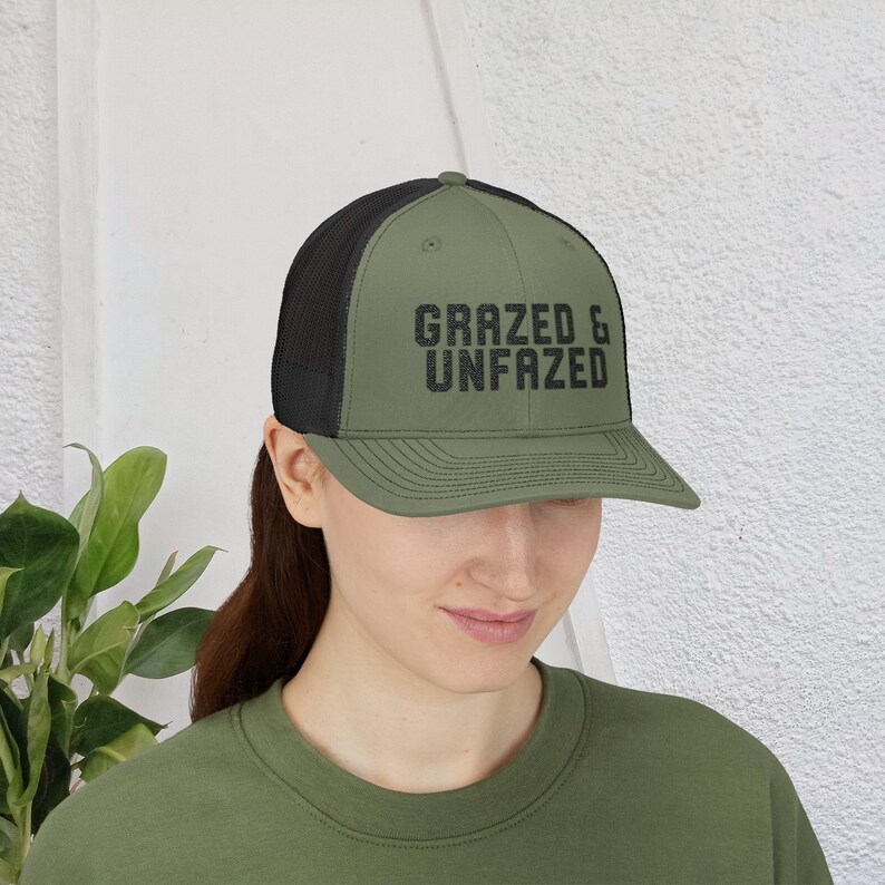 May include: Olive green trucker hat with a black mesh back. The hat features the text "GRAZED & UNFAZED" in a distressed black font across the front. The hat is worn by a person.