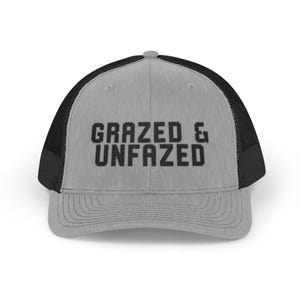 May include: A gray trucker hat with a black mesh back and a curved brim. The front of the hat features the text "GRAZED & UNFAZED" in bold, black letters. The hat is designed to be worn by anyone.