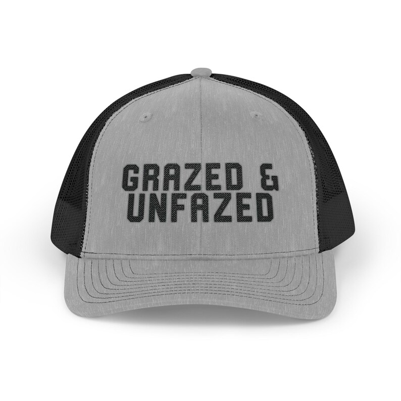 May include: A gray trucker hat with a black mesh back and a curved brim. The front of the hat features the text "GRAZED & UNFAZED" in bold, black letters. The hat is designed to be worn by anyone.