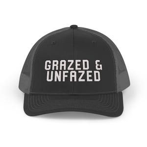 May include: A black trucker hat with a mesh back and a curved brim. The front of the hat features the white text "GRAZED & UNFAZED" in a bold, sans-serif font. The hat is designed for casual wear.
