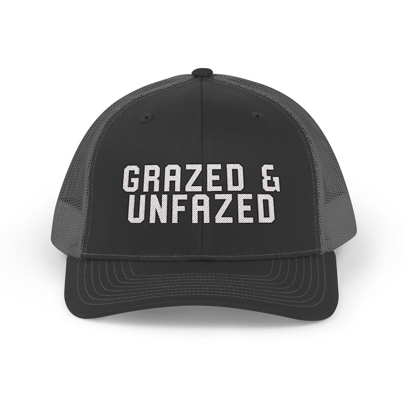 May include: A black trucker hat with a mesh back and a curved brim. The front of the hat features the white text "GRAZED & UNFAZED" in a bold, sans-serif font. The hat is designed for casual wear.